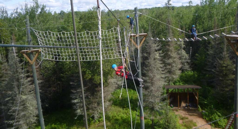 On a high ropes course, three people wearing safety gear and secured by ropes make their way through the course. 